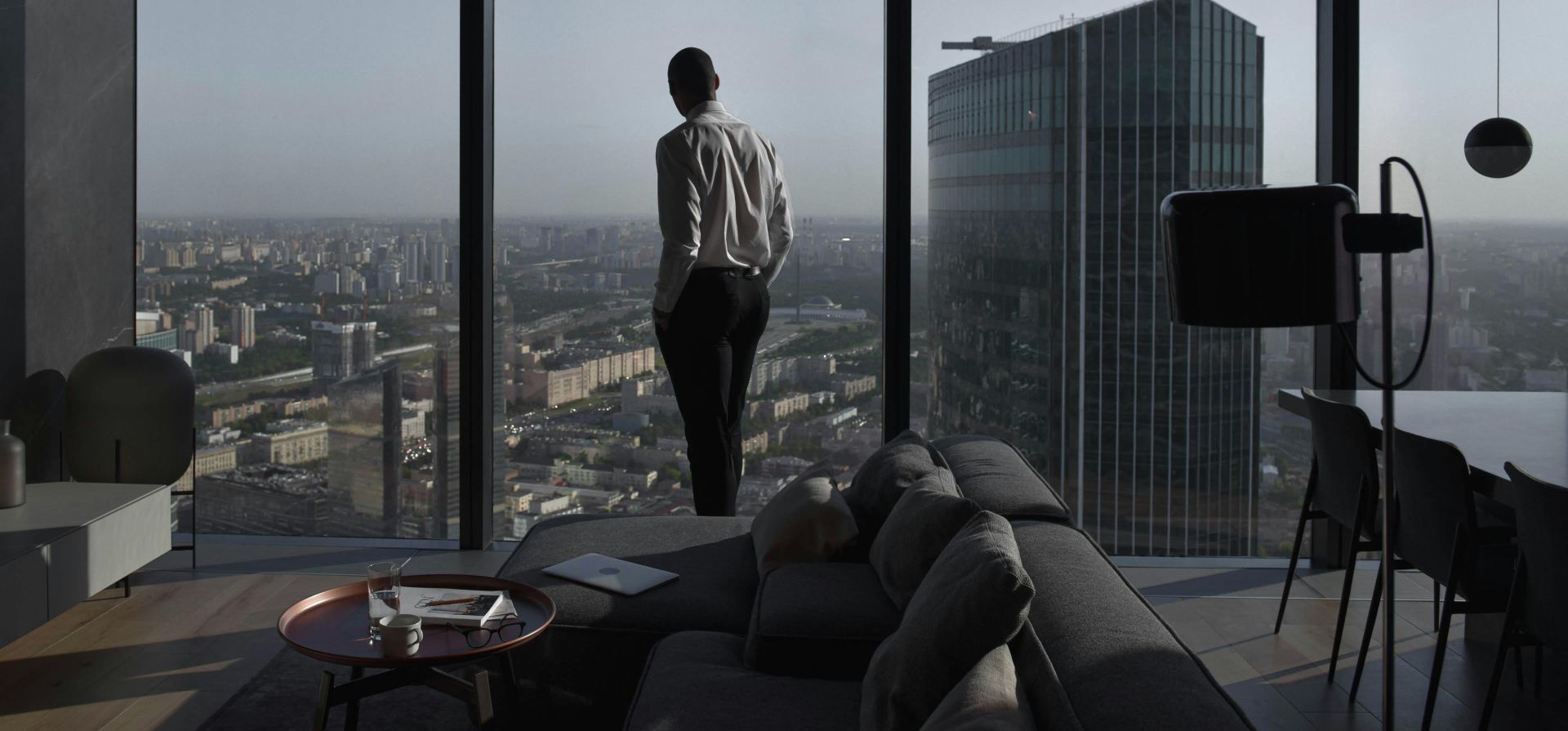 Man in a modern apartment with a panoramic view of an urban skyline and skyscraper.